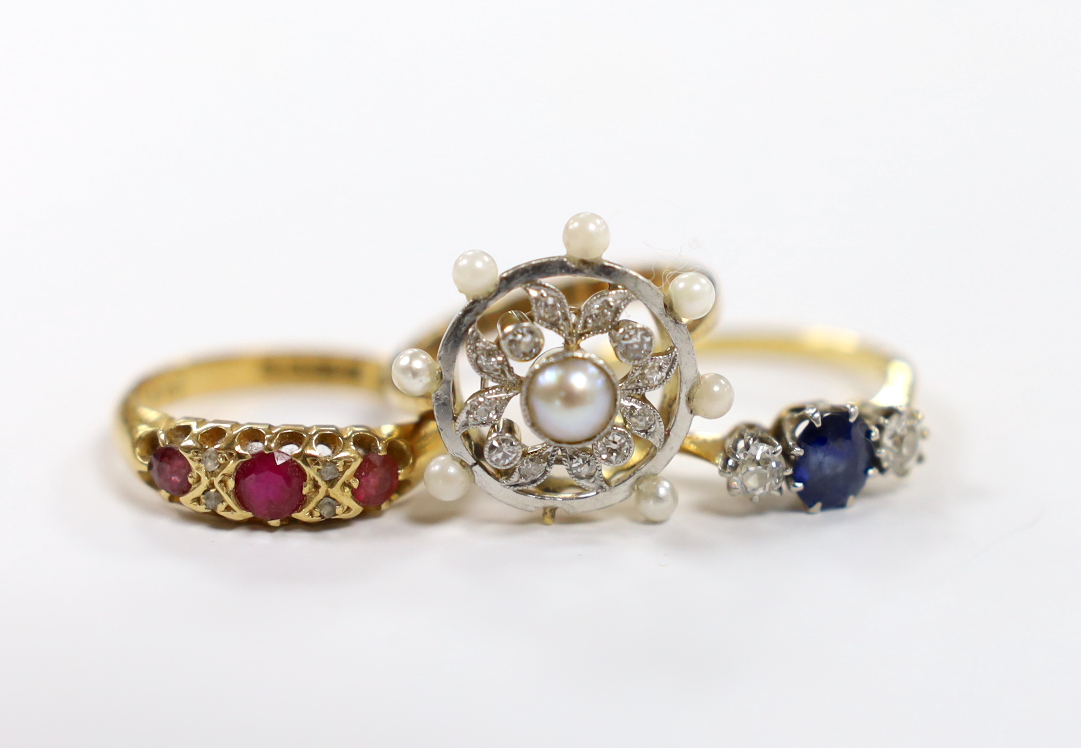 An 18ct, sapphire and diamond set three stone ring, a George V 18ct gold and three red stone set ring with diamond chip spacers and a yellow metal seed pearl and diamond chip set cluster ring (pearl missing), gross weigh